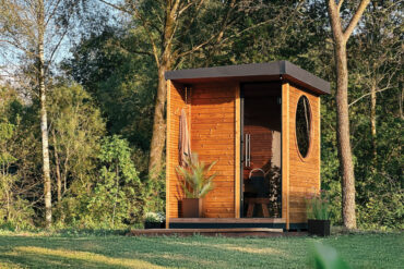 Auroom Kaia Außensauna mit Thermory Thermo-Fichte Wandverkleidung und rundem Fenster, umgeben von grünem Wald.