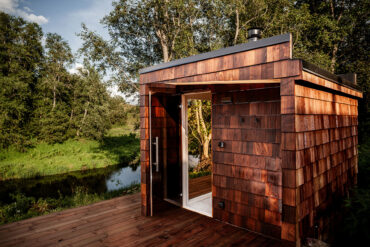 Jõeäärne Thermory termosaare sindlitega kaetud saun, mida ümbritseb rohelus ja looduslik valgus, rõhutades puidupinna tekstuuri.