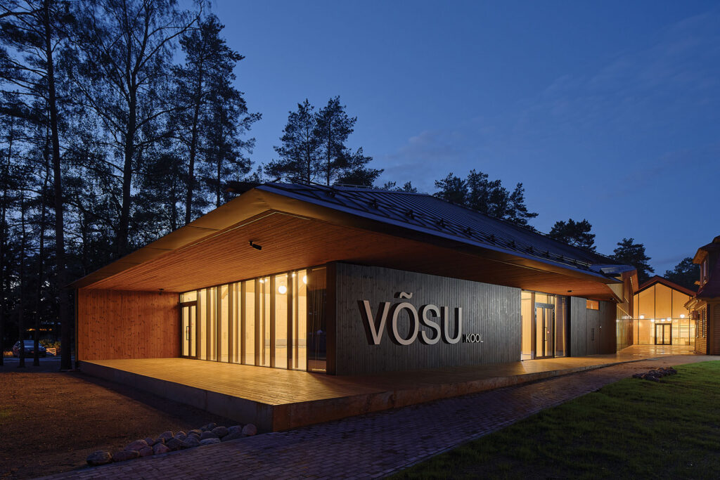 Evening exterior of Võsu School showing dark brushed Thermory thermo-pine cladding and warm-lit interiors among pine trees.